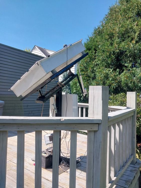 Solar panel mounted on a deck, angled towards the sun, in front of a house and trees.