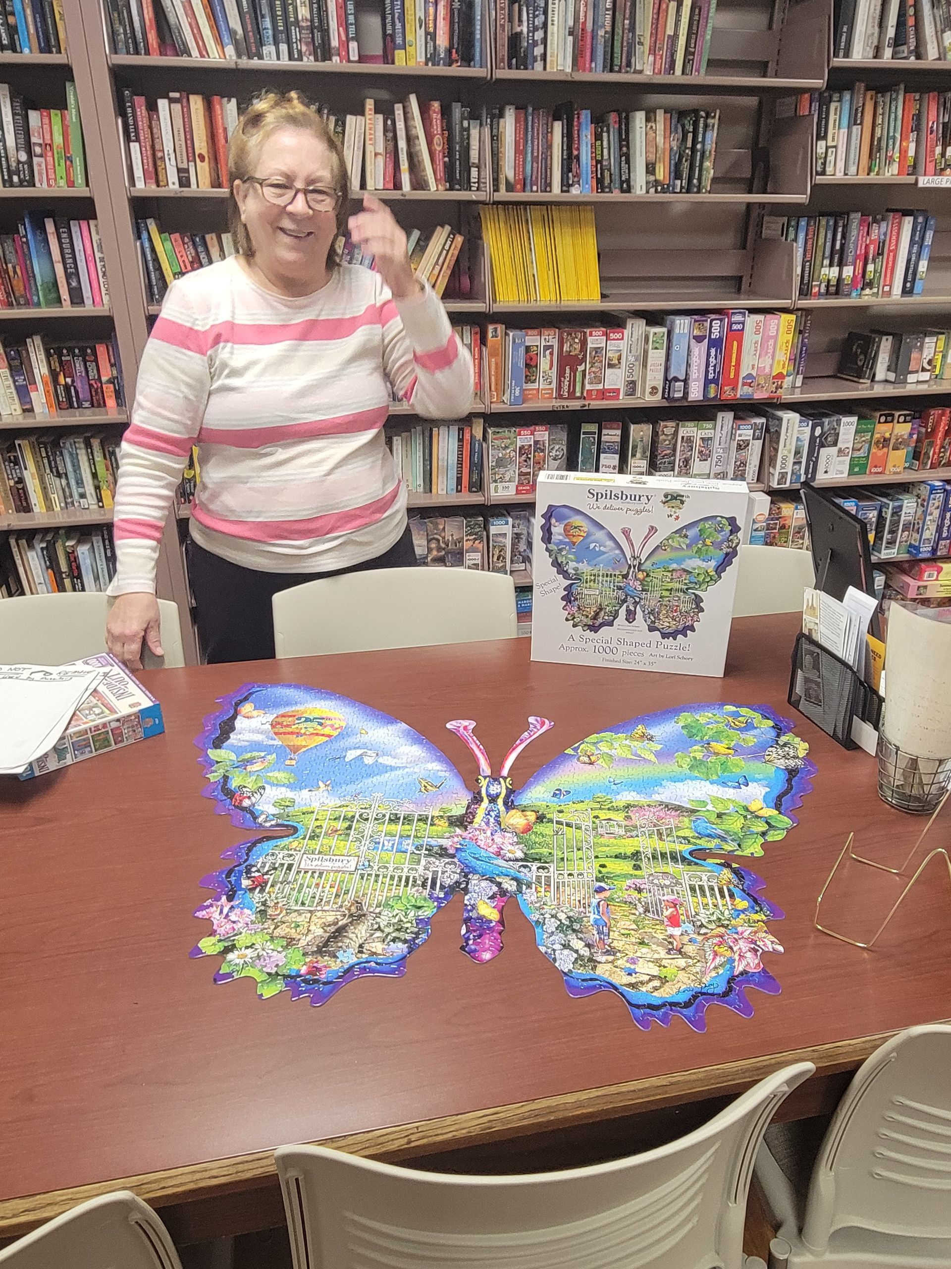 A person smiles near a table with a butterfly-shaped jigsaw puzzle and its box in a library with bookshelf backgrounds.