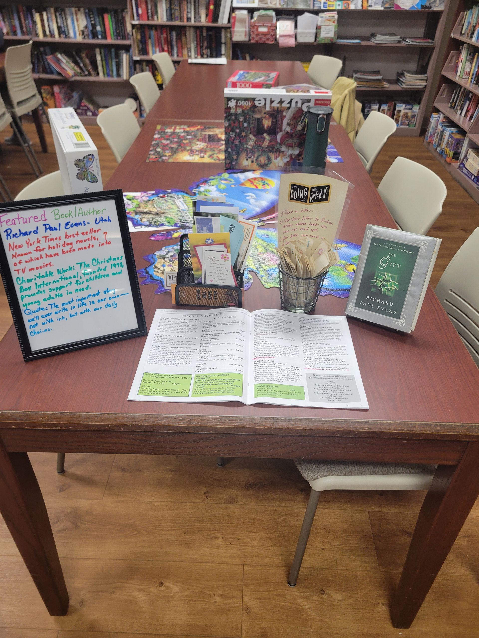 A library table set up for a game activity, with a sign, board game boxes, a tablet, and a worksheet with green notes.