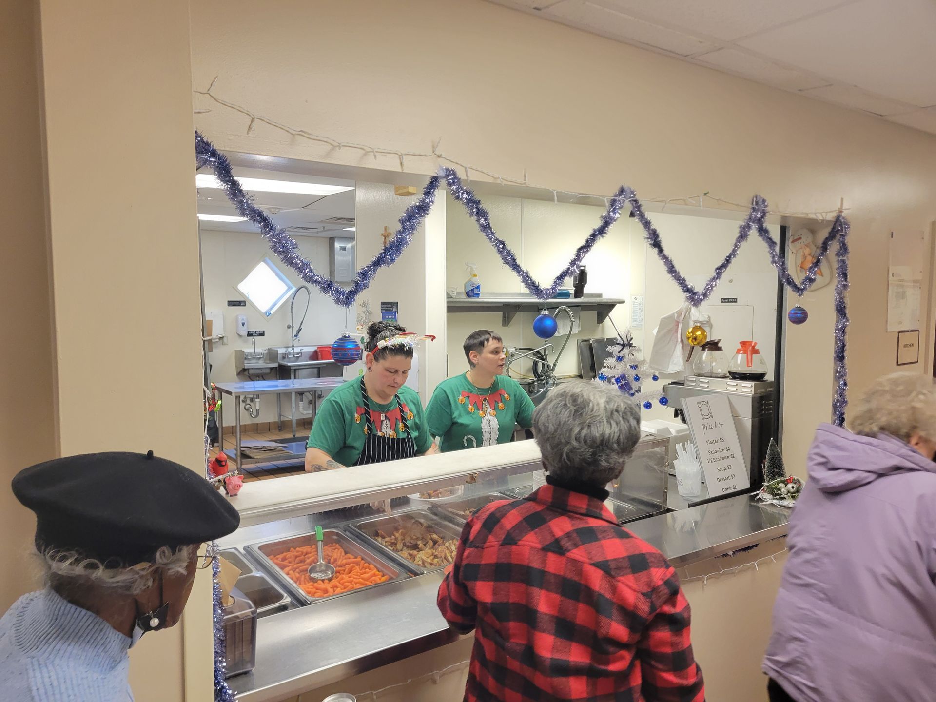 People stand in a buffet line in a room decorated with blue tinsel and ornaments for a holiday meal service.