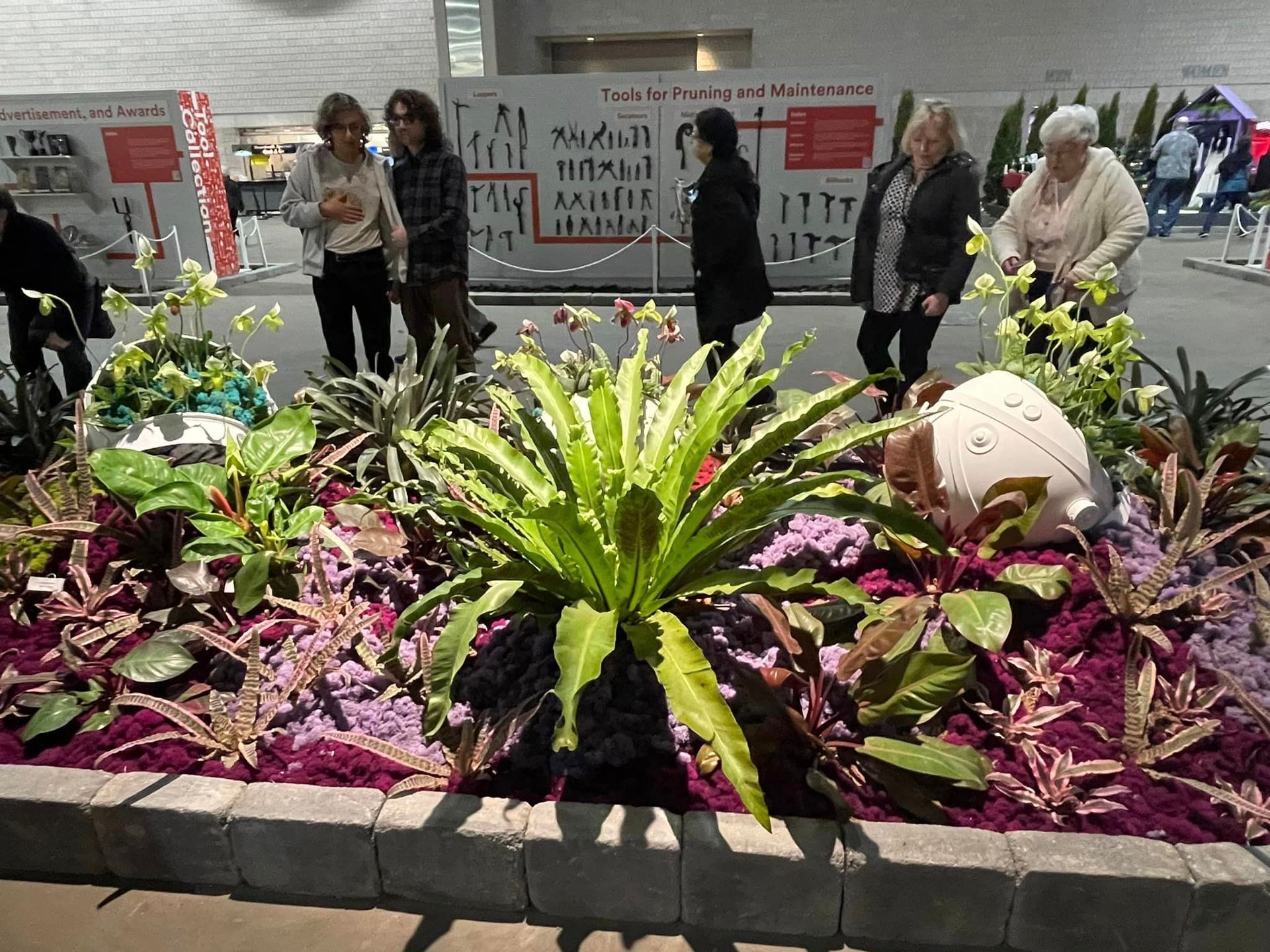 A vibrant floral exhibit with a large central fern, surrounded by purple ground cover and plants, viewed in an atrium.