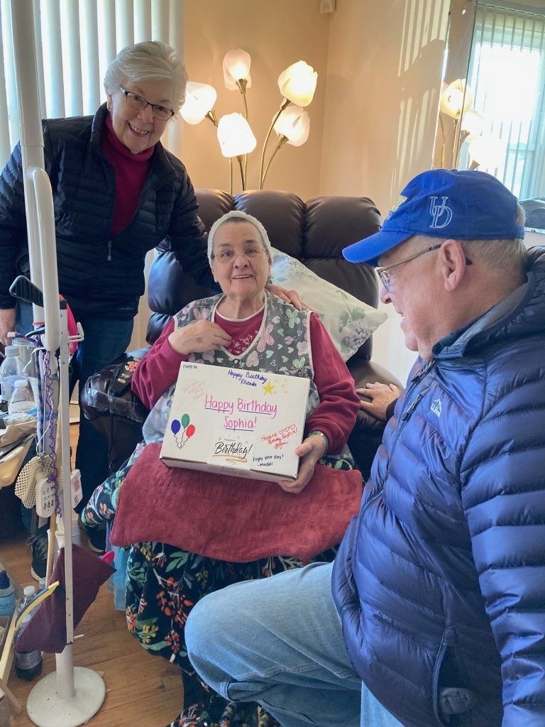 A person sitting in a recliner holding a decorated gift box, flanked by two people, one standing and one kneeling nearby.
