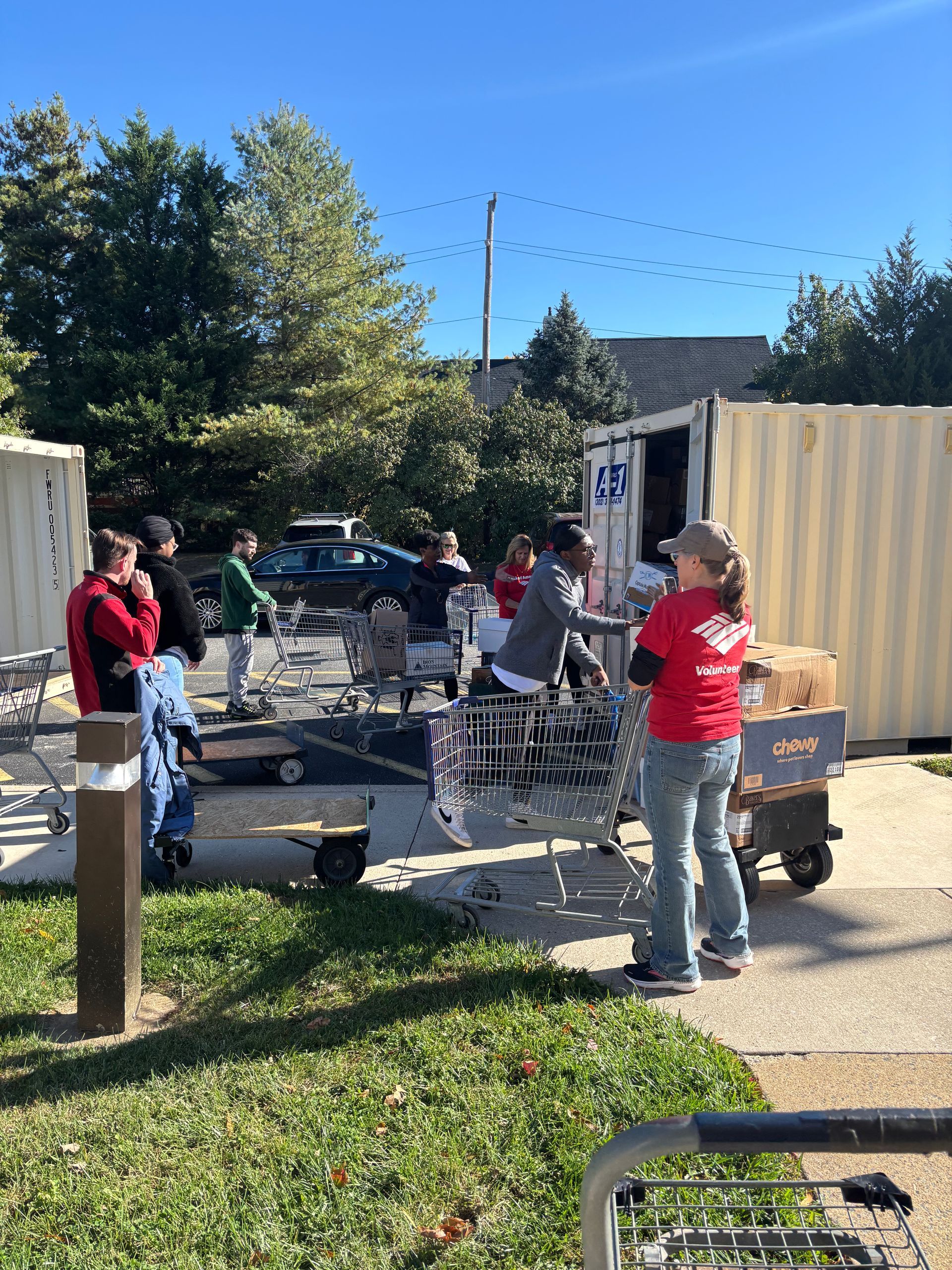 Volunteers in casual clothing load groceries into shopping carts outside a shipping container on a sunny day.