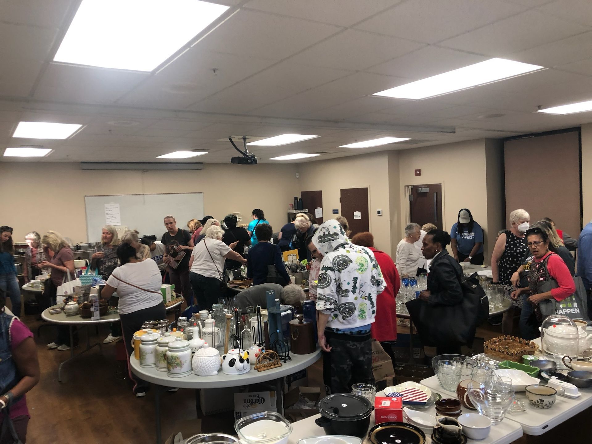 People browse a variety of household goods and ceramics displayed on tables at an indoor rummage sale.