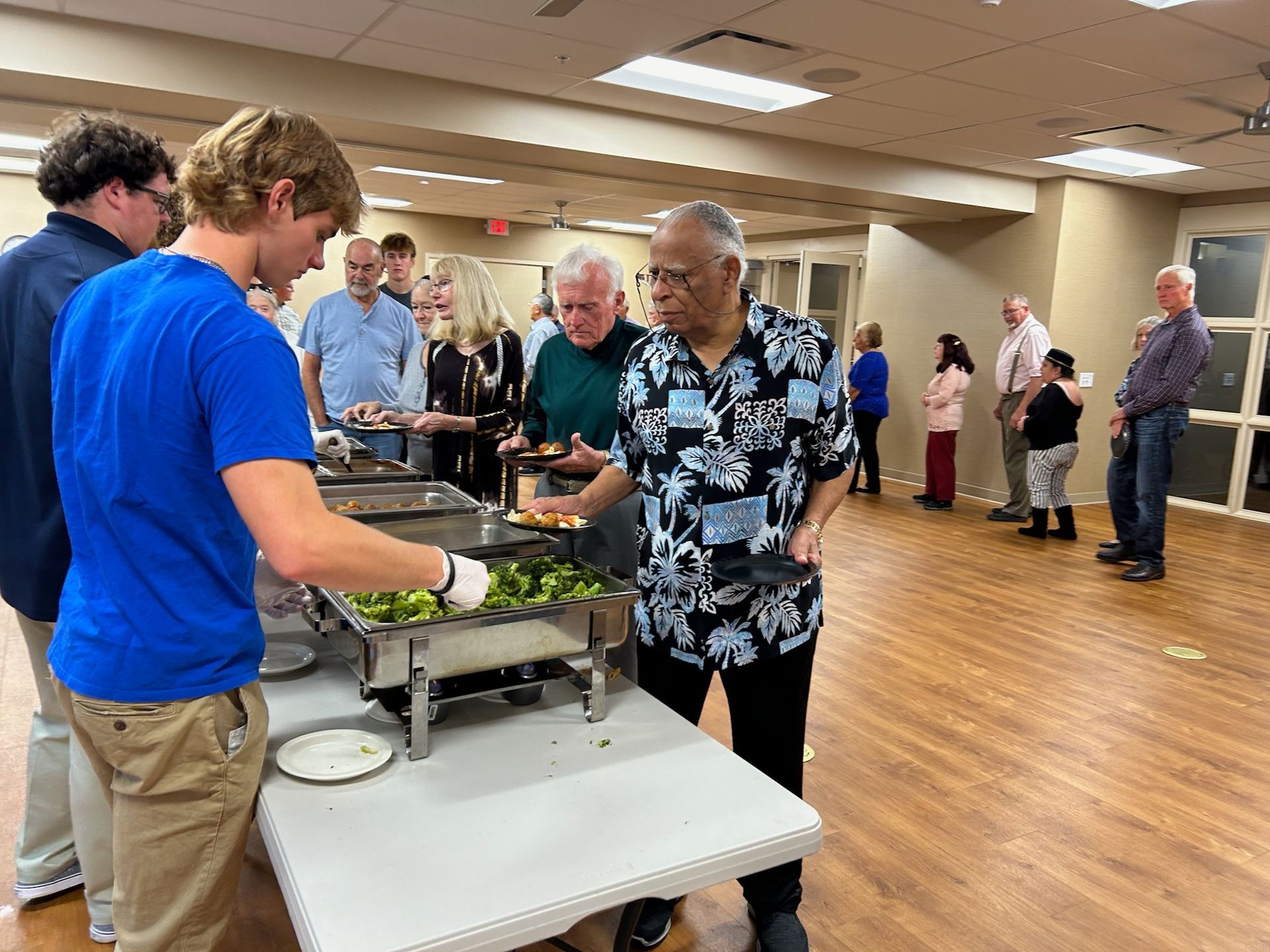 People in a community hall serve themselves from a buffet line with trays of food set out on long tables.