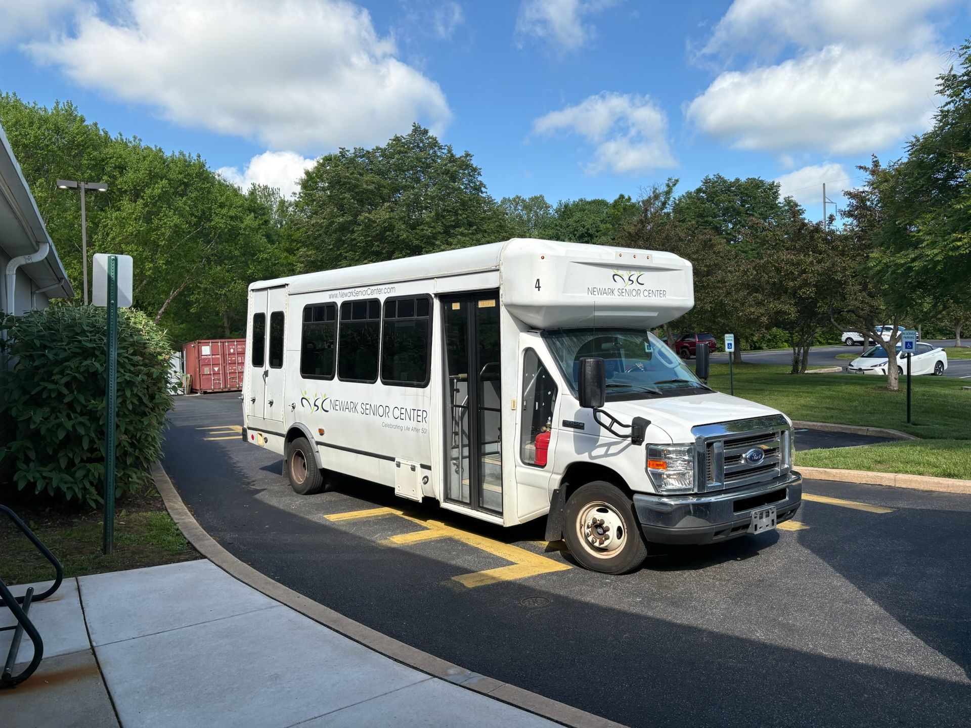 A white shuttle bus parked in a paved lot on a sunny day with green trees in the background.