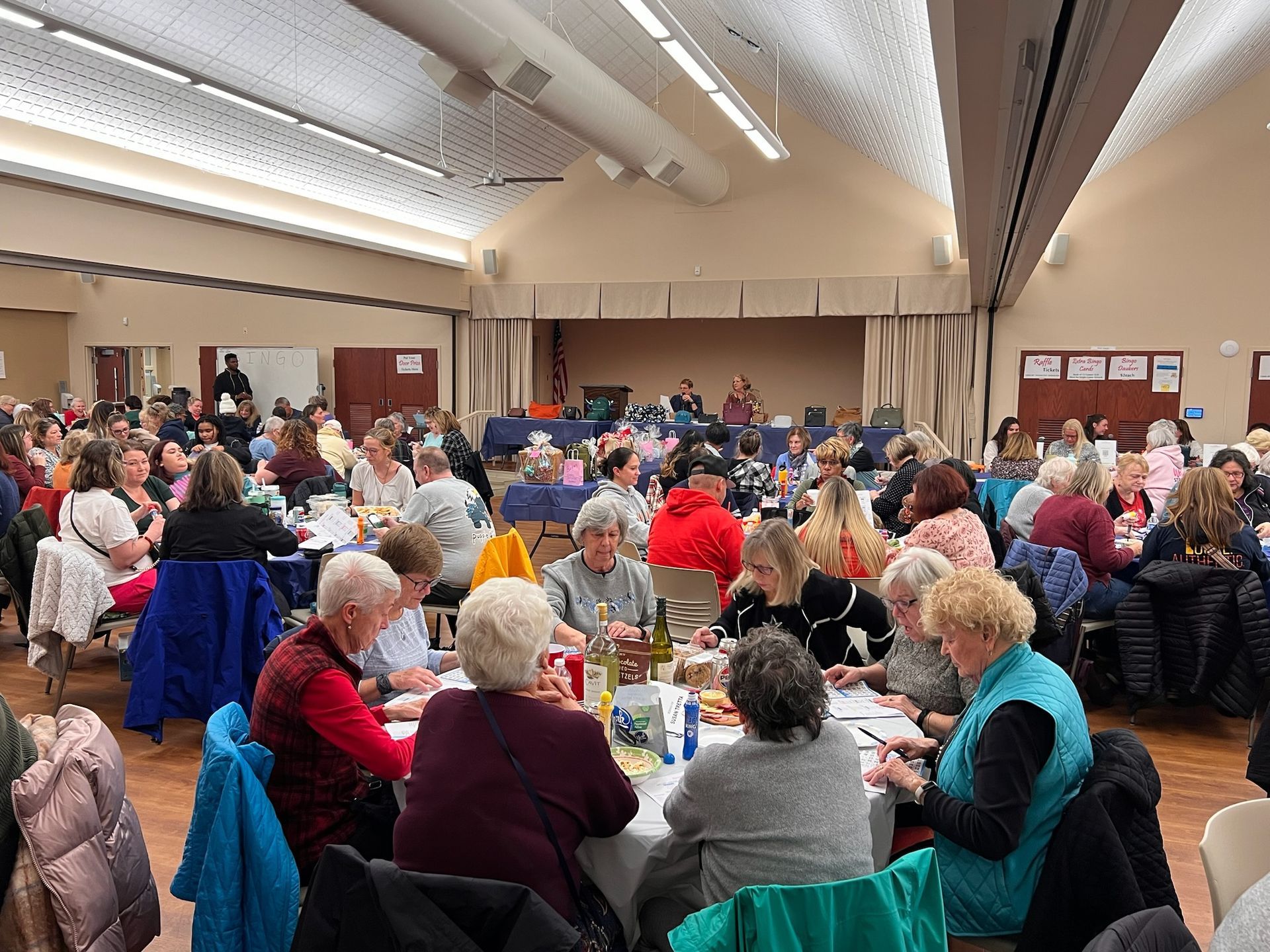 A large group of people seated at round tables in a spacious hall for a social gathering or meal, with a stage at the back.