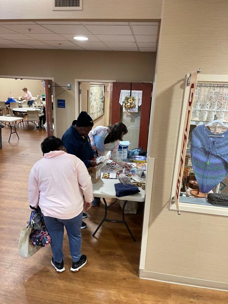 People stand at a table in an indoor hallway, interacting with items laid out for display or distribution.