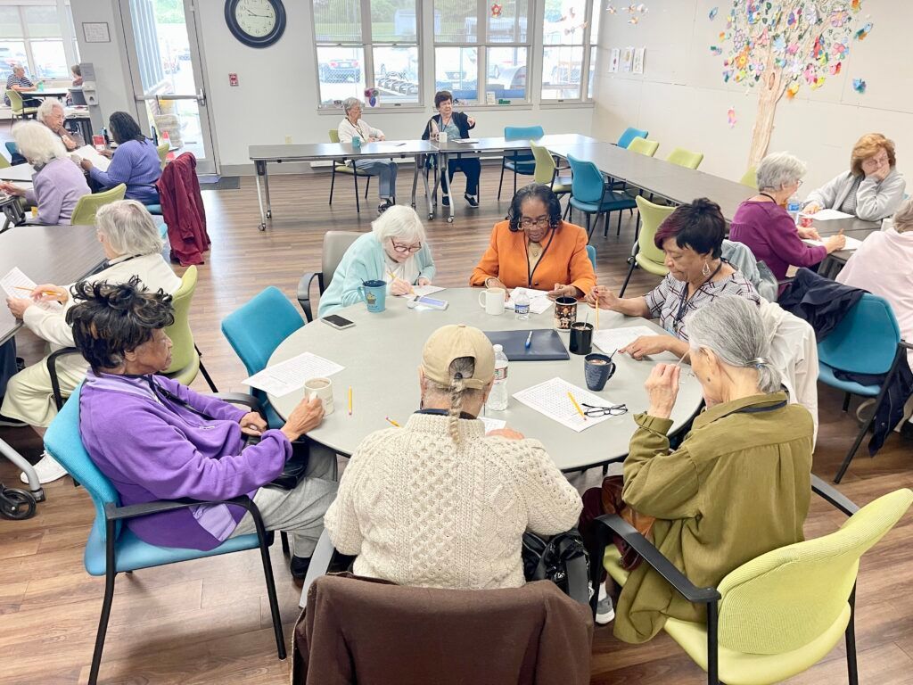 A group of people sit around a circular table in a bright community room, working together on papers and tasks.