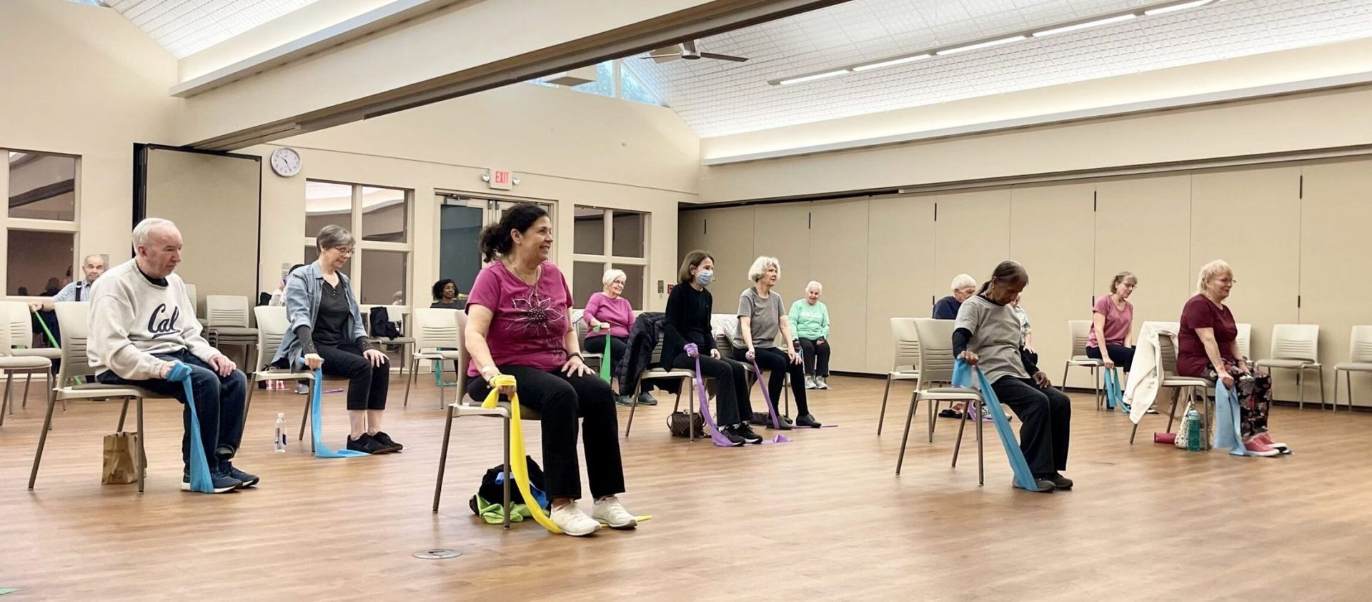 A group of people sit in a line of chairs in a studio, performing seated exercises using colorful resistance bands.