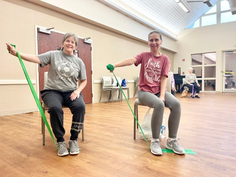Two people seated in a studio exercise with green resistance bands held in one hand and anchored under their feet.