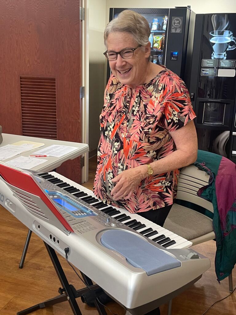 A smiling person in a floral shirt plays a keyboard on a stand in a room with a vending machine.