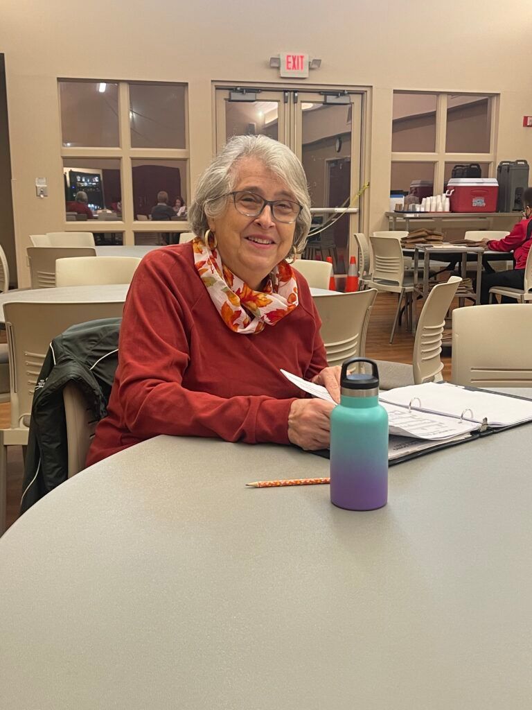 A person wearing a red sweater and colorful scarf sits at a table, writing in a binder with a water bottle nearby.