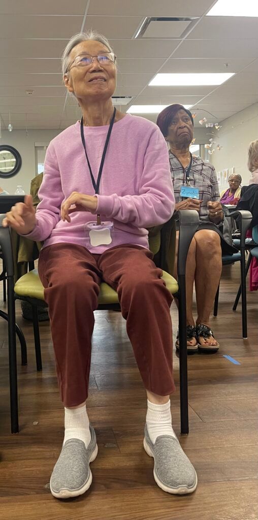 A person in a light purple sweater and maroon pants sits in a chair in a room, looking upward with their hand raised.