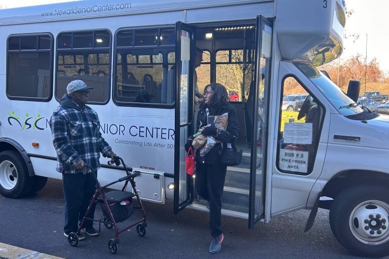 A person with a walker stands outside a white bus labeled