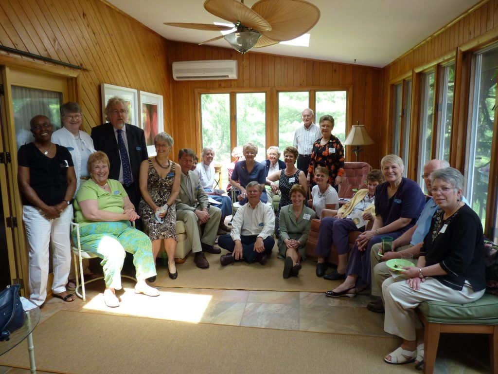 A group of people posed in a wood-paneled sunroom with large windows, some seated and some standing together for a photo.