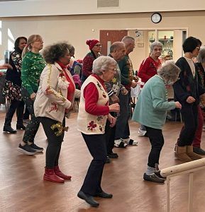 A group of people wearing festive clothing participate in a line dance in a bright community room.