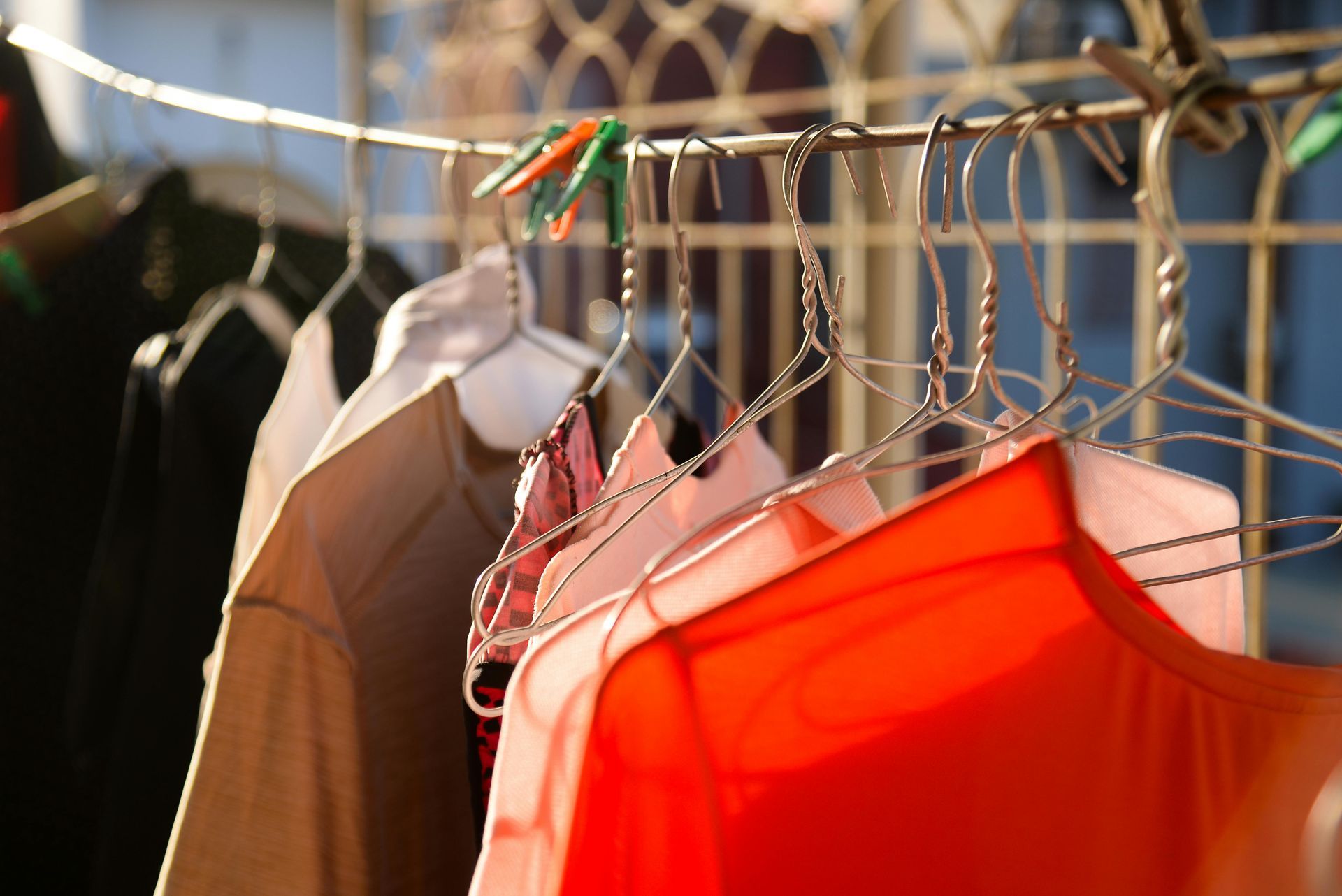 Several t-shirts in shades of orange, pink, and black hang on clothes hangers on a line outside.