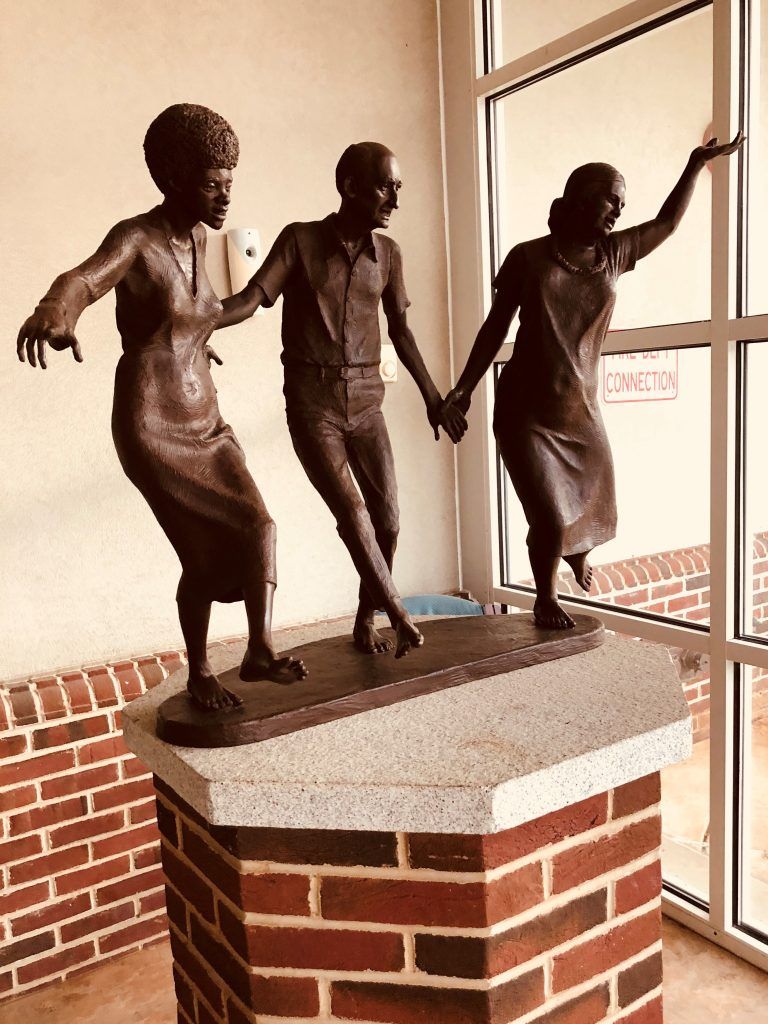 A bronze sculpture of three figures holding hands in a dance pose, mounted on a brick and stone pedestal near a window.