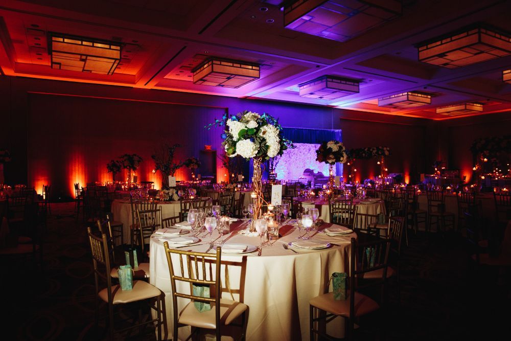 A dimly lit banquet hall features round tables with white tablecloths, floral centerpieces, and warm orange and blue light.