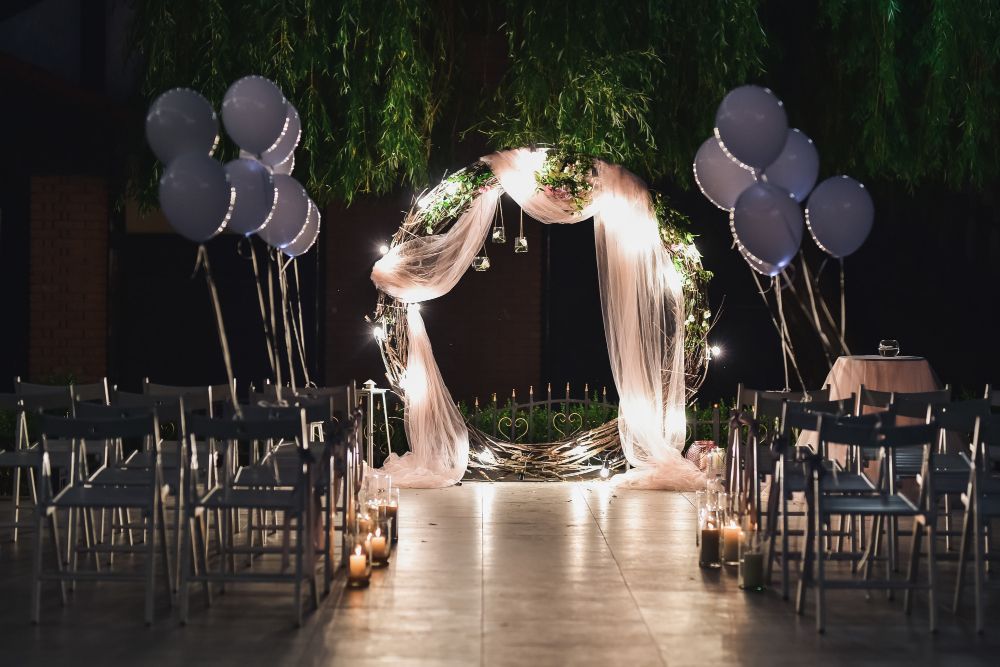 A wedding ceremony setup featuring a circular floral arch with white drapes, fairy lights, and floating balloons at night.