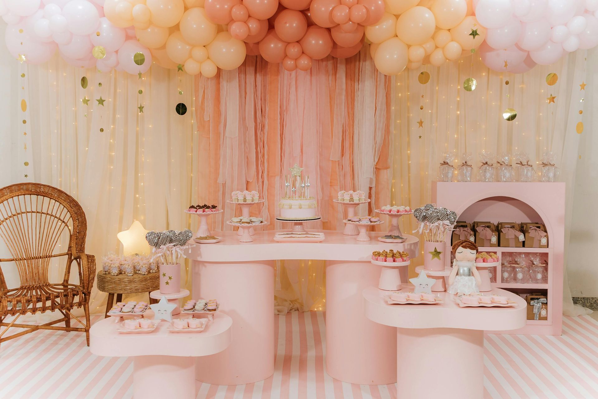 A party setup with a pink balloon arch, tiered tables, a rattan chair, and assorted desserts in a pastel color scheme.