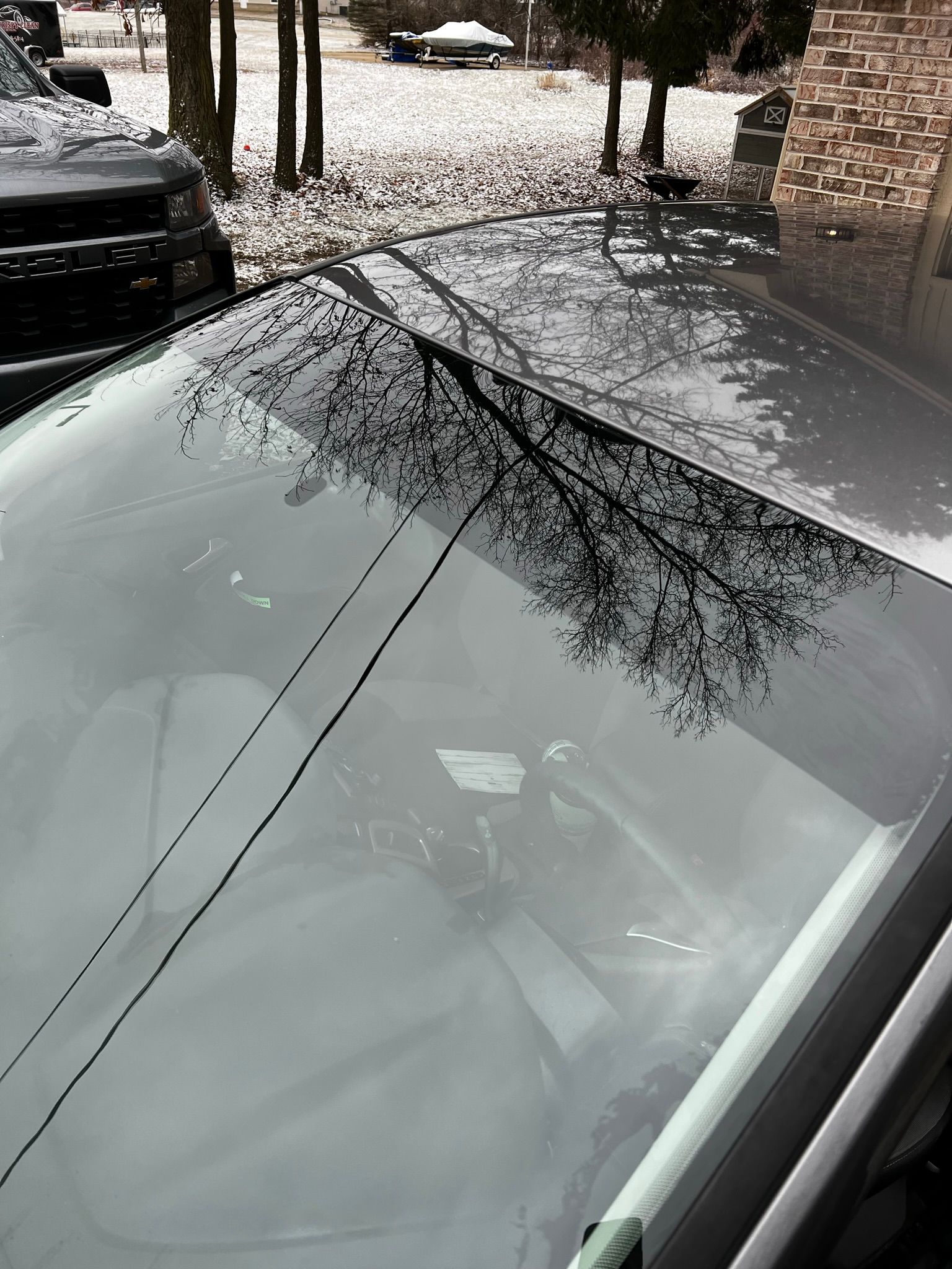A car is parked in the snow with a reflection of trees in the windshield.