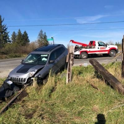 Gray SUV crashed off road next to a wooden fence. A tow truck is present, clear blue sky.