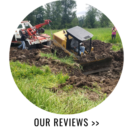 A bulldozer stuck in mud being pulled out by a tow truck; two people stand nearby.