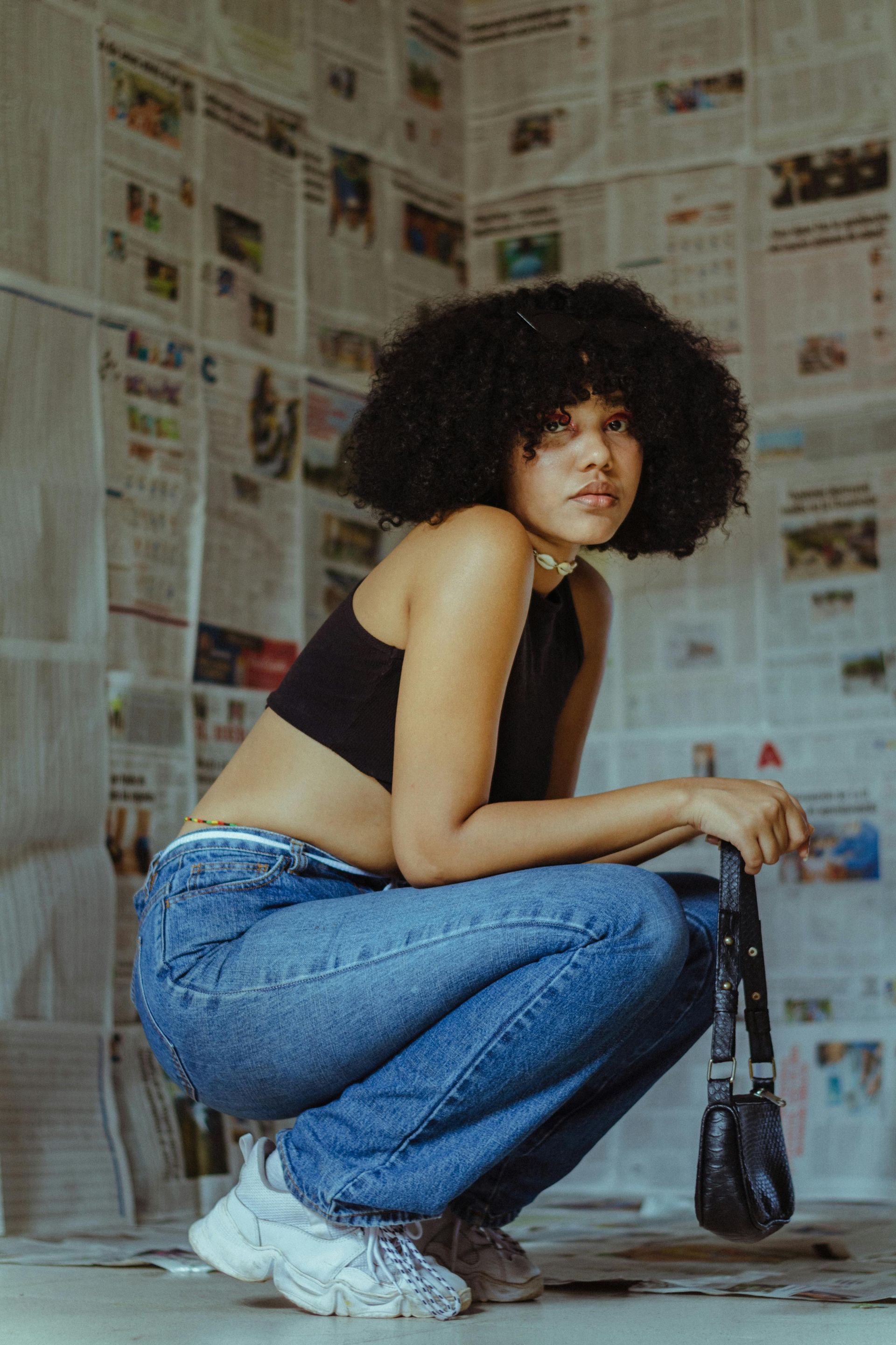 A woman with curly hair is squatting down in front of a wall of newspapers.