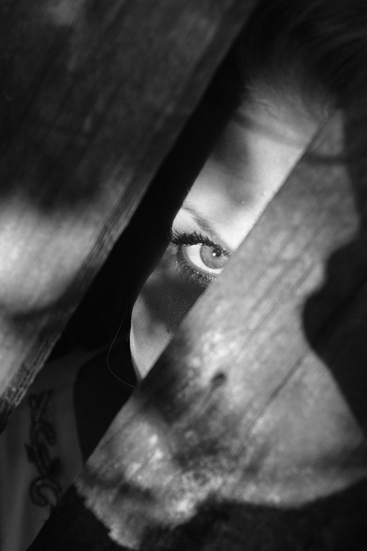 A black and white photo of a woman 's eye peeking out from behind a wooden fence.