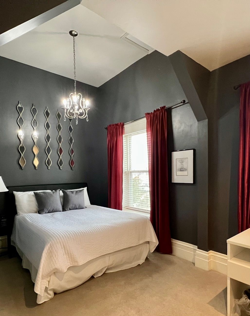 Dark gray bedroom with red curtains, chandelier, decorative wall mirrors, and bed.