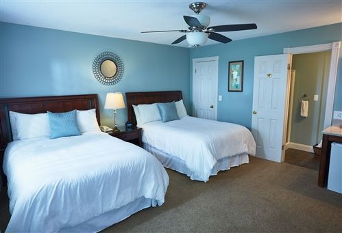 Two beds with white linens in a light blue hotel room, with a ceiling fan and a doorway to a bathroom.