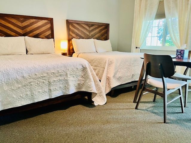 Two double beds in a hotel room with wooden headboards, a window, and a small table with chairs.