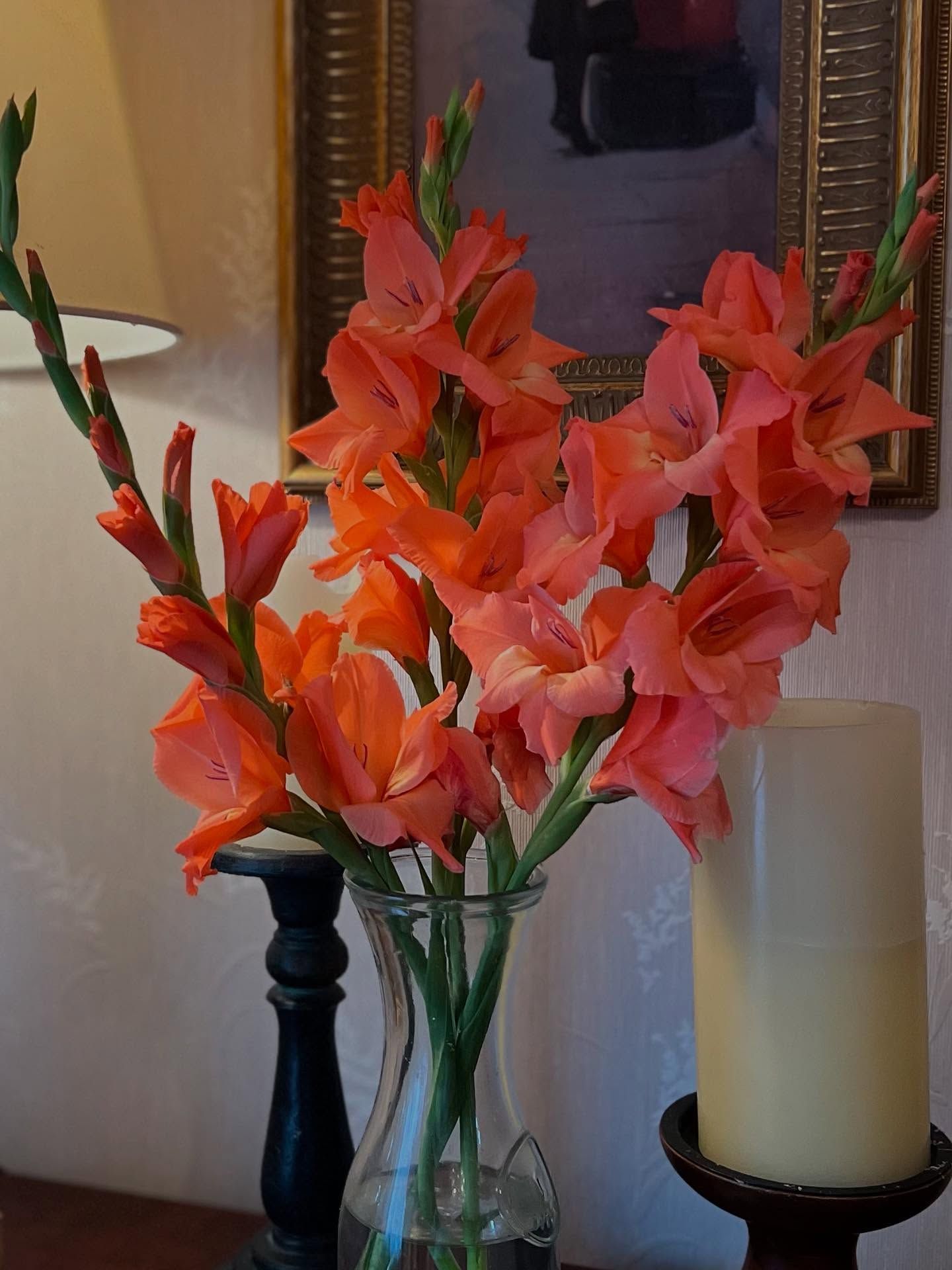 Orange gladiolus flowers in a glass vase, with a candle and framed art in the background.