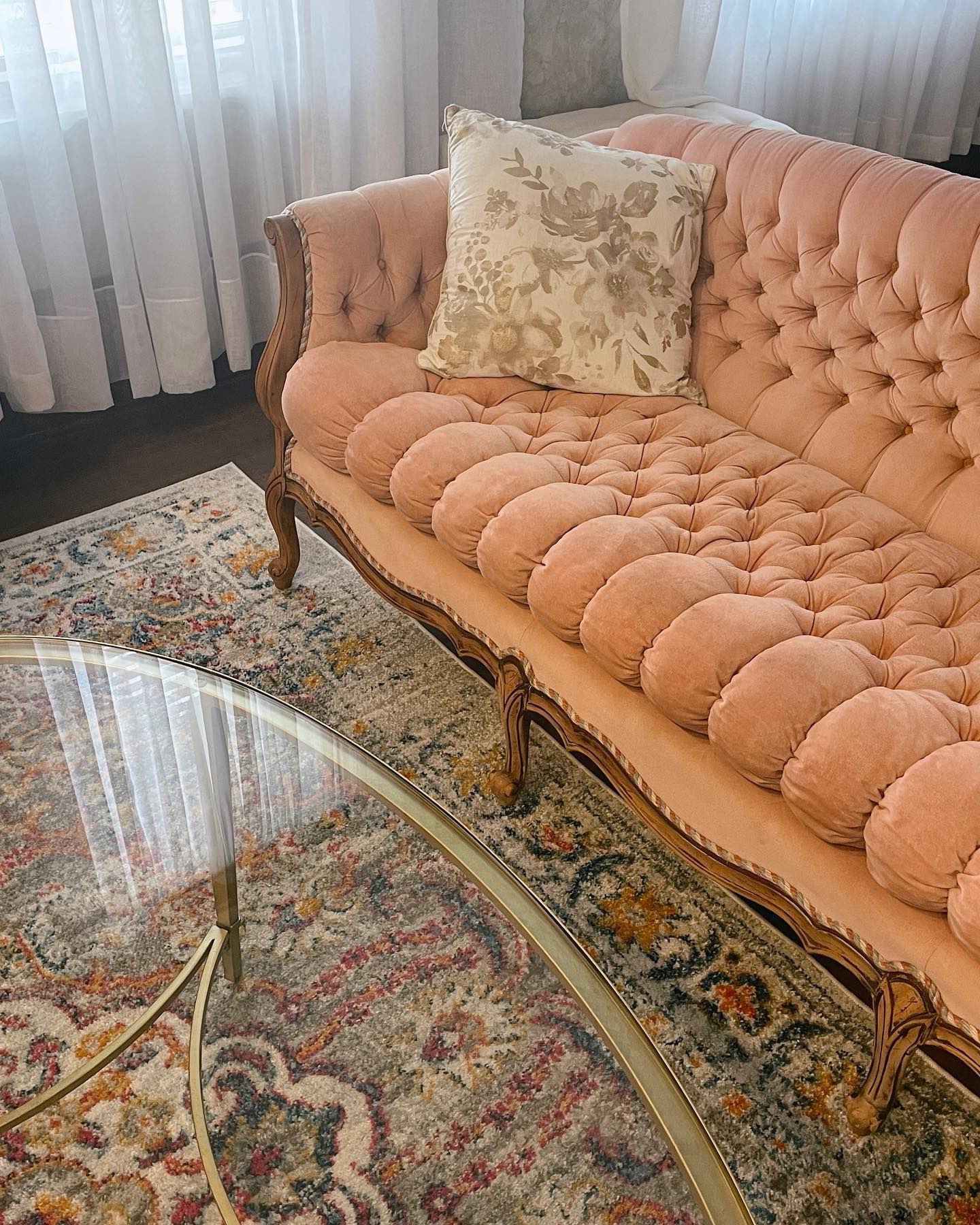 Pink tufted sofa with floral pillow, ornate wooden frame. Glass and gold coffee table, patterned rug.