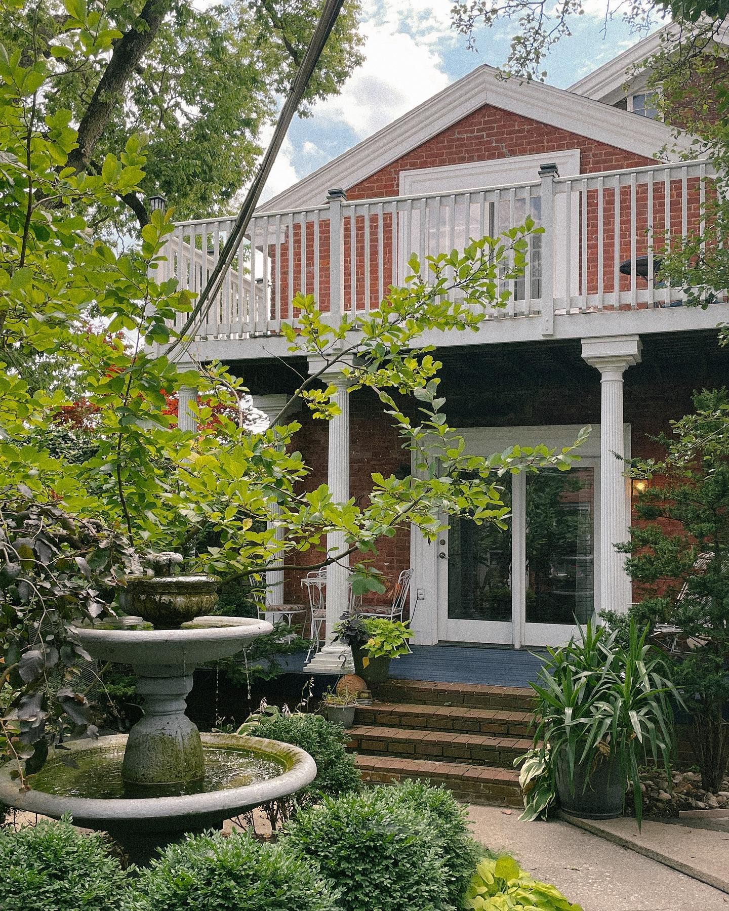Brick building with white balcony, fountain, and lush greenery.