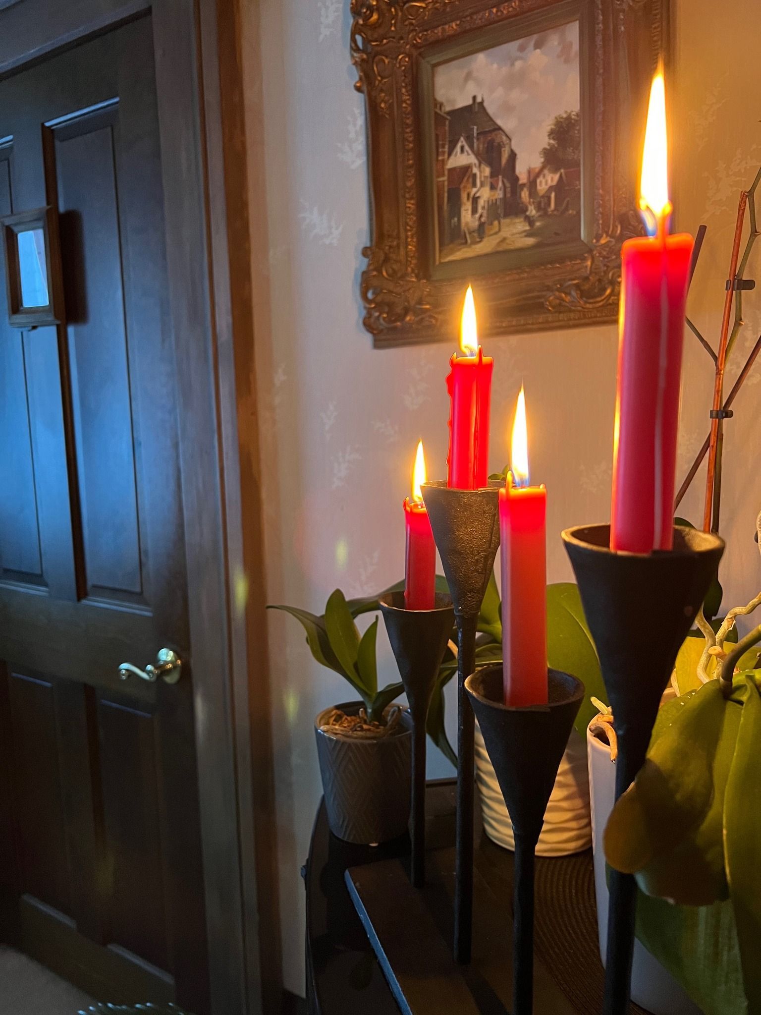 Burning red candles on black stands, next to a door, beneath a painting.