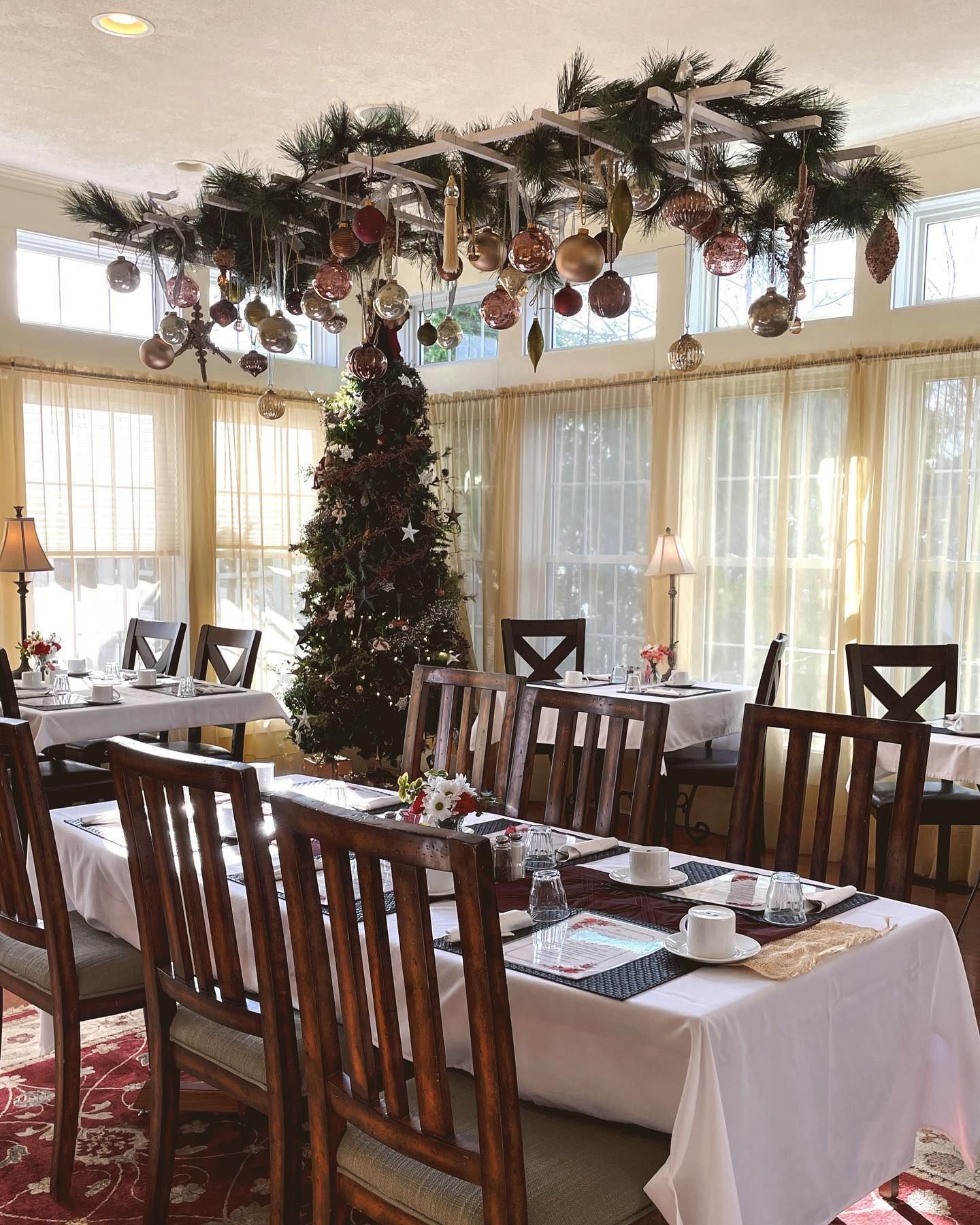 Dining room decorated for Christmas with a tree, ornaments, tables set for a meal, and sheer curtains.
