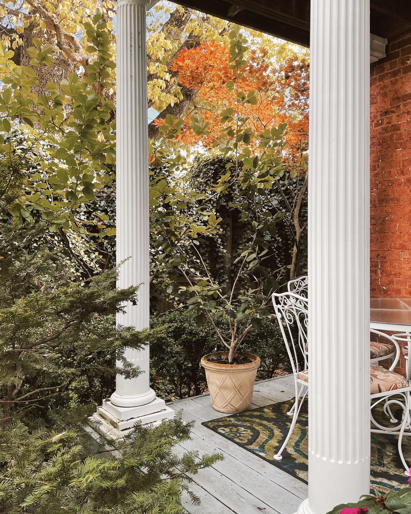 White columns frame a porch with a small potted plant and wrought iron furniture, surrounded by autumn foliage.