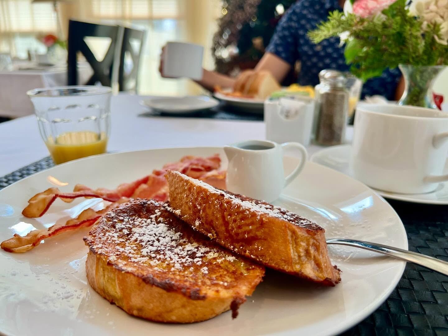 Breakfast plate with French toast, bacon, syrup, orange juice, and a coffee cup on a table.