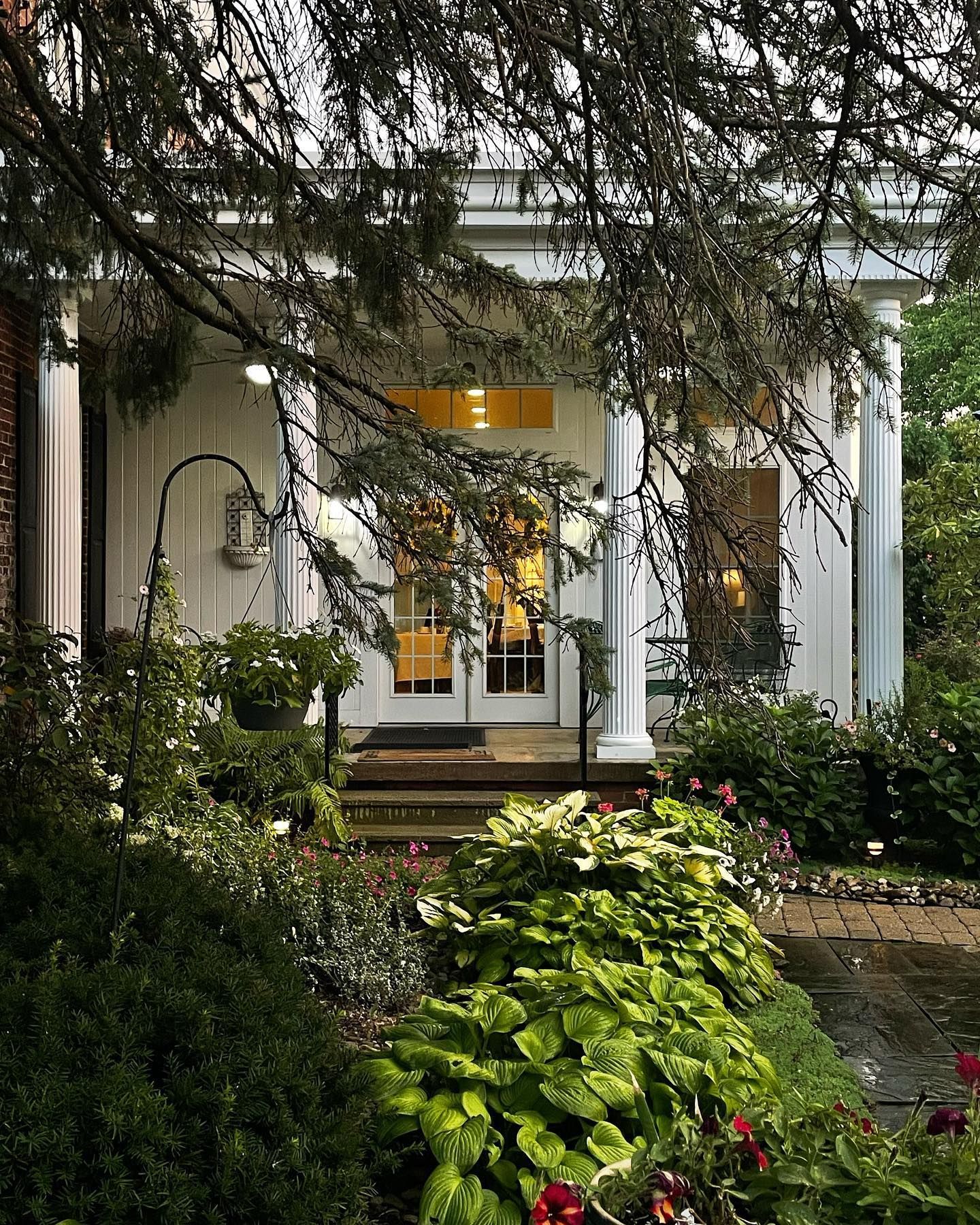 White-columned building entrance with a lush garden; warm light glows from within.