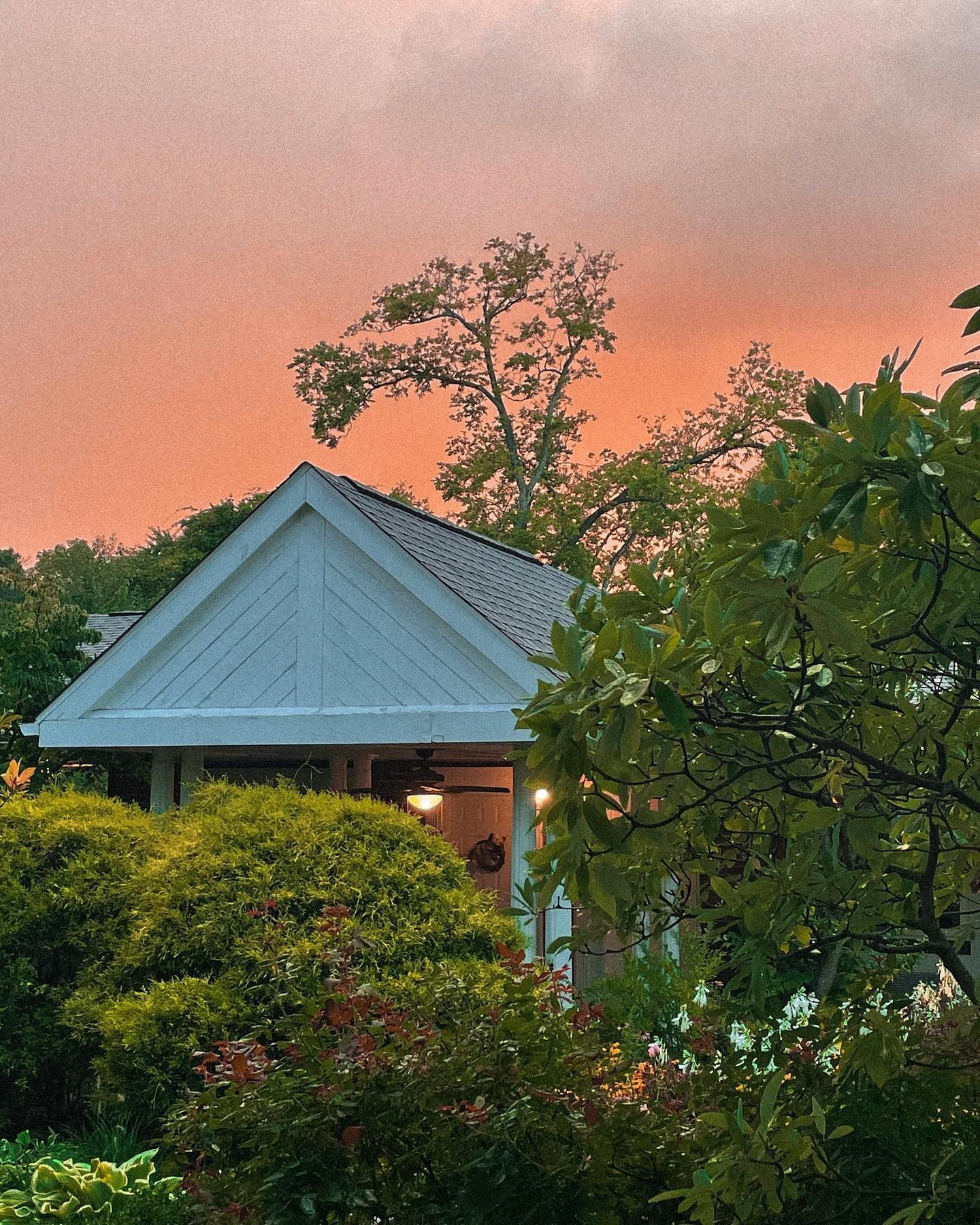 White-roofed house with porch, surrounded by green foliage, under an orange and pink sunset sky.