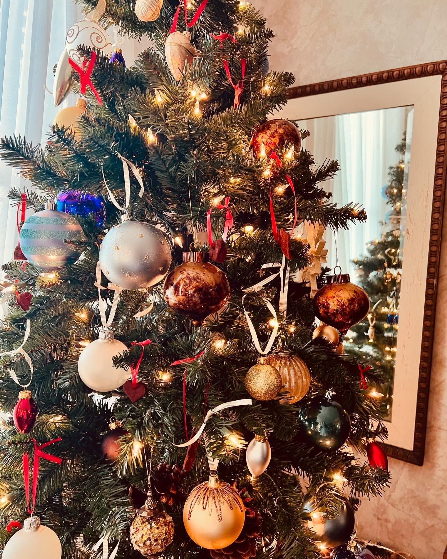 Christmas tree decorated with ornaments, fairy lights, and a mirror in the background reflecting another tree.