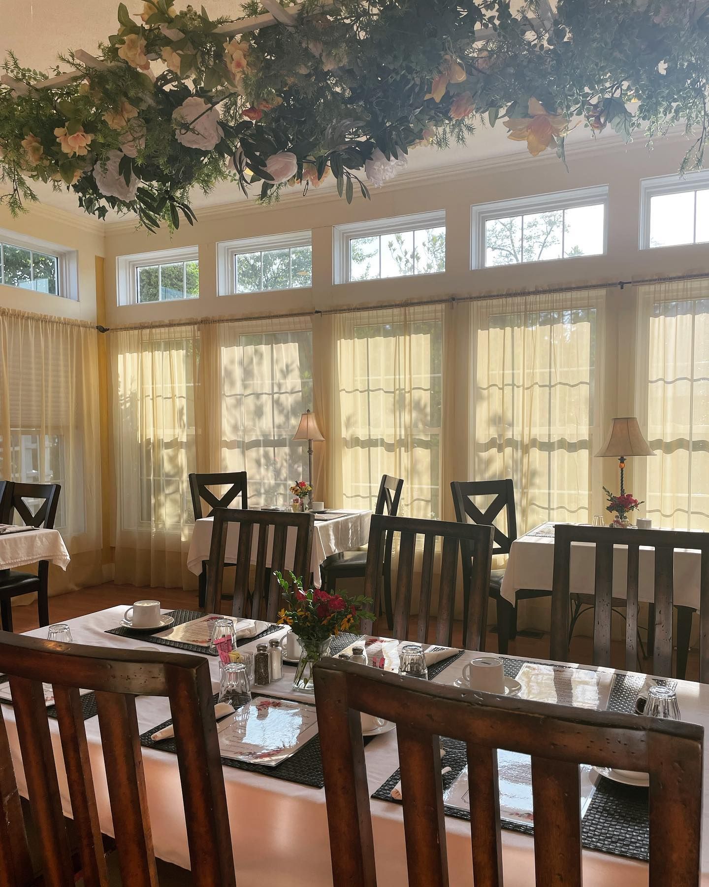 Dining room with tables set for a meal; floral decorations hang from the ceiling. Yellow curtains and sunlight.