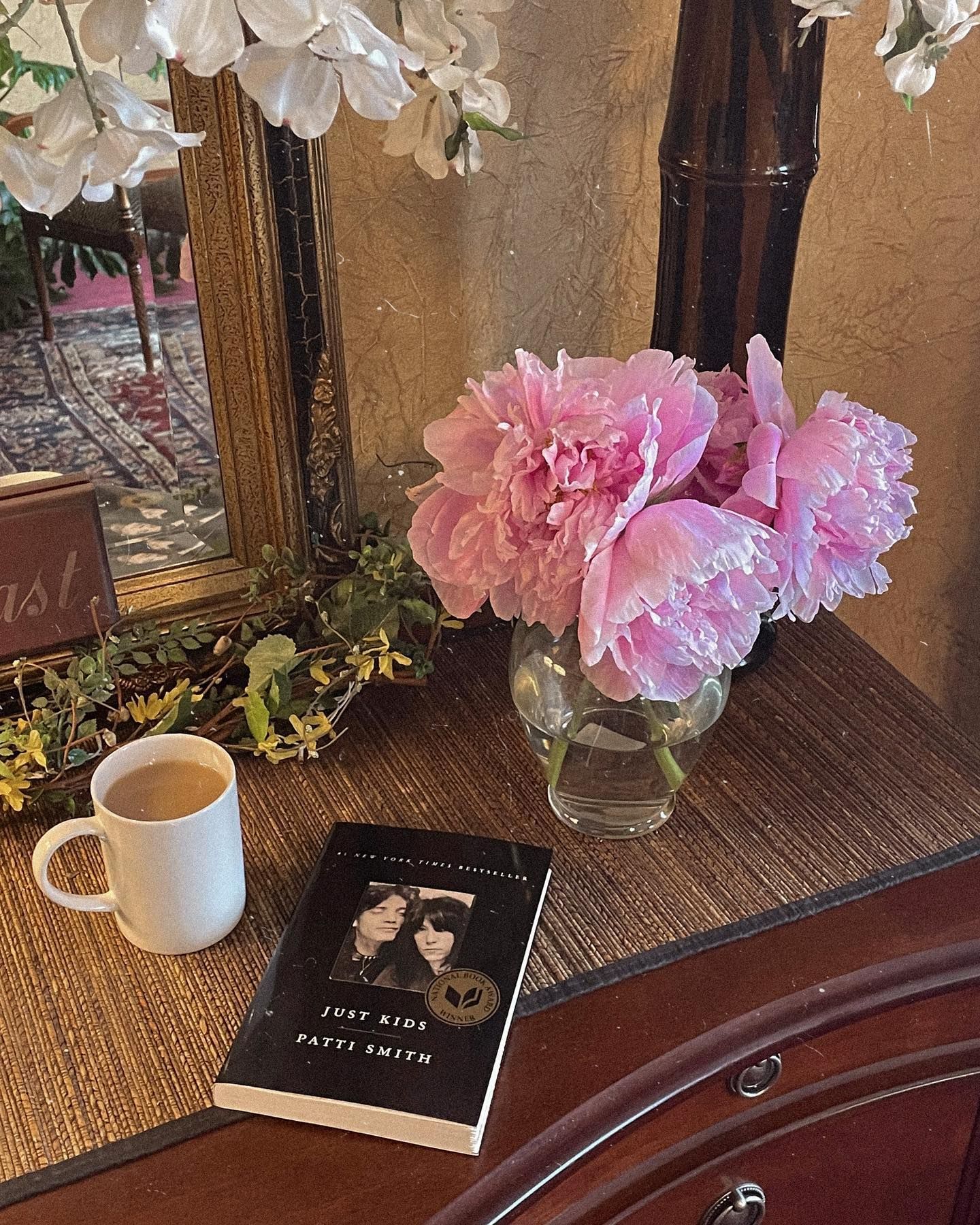 Pink peonies in a vase, coffee mug, and a book on a wooden table. Mirror in the background.