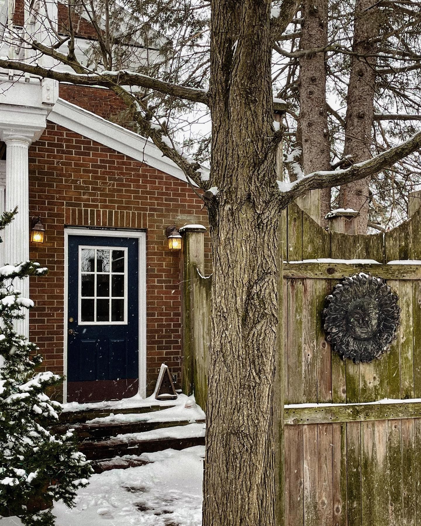 Snowy brick building with a navy door and a wooden fence with a wreath. A tree trunk is in the foreground.