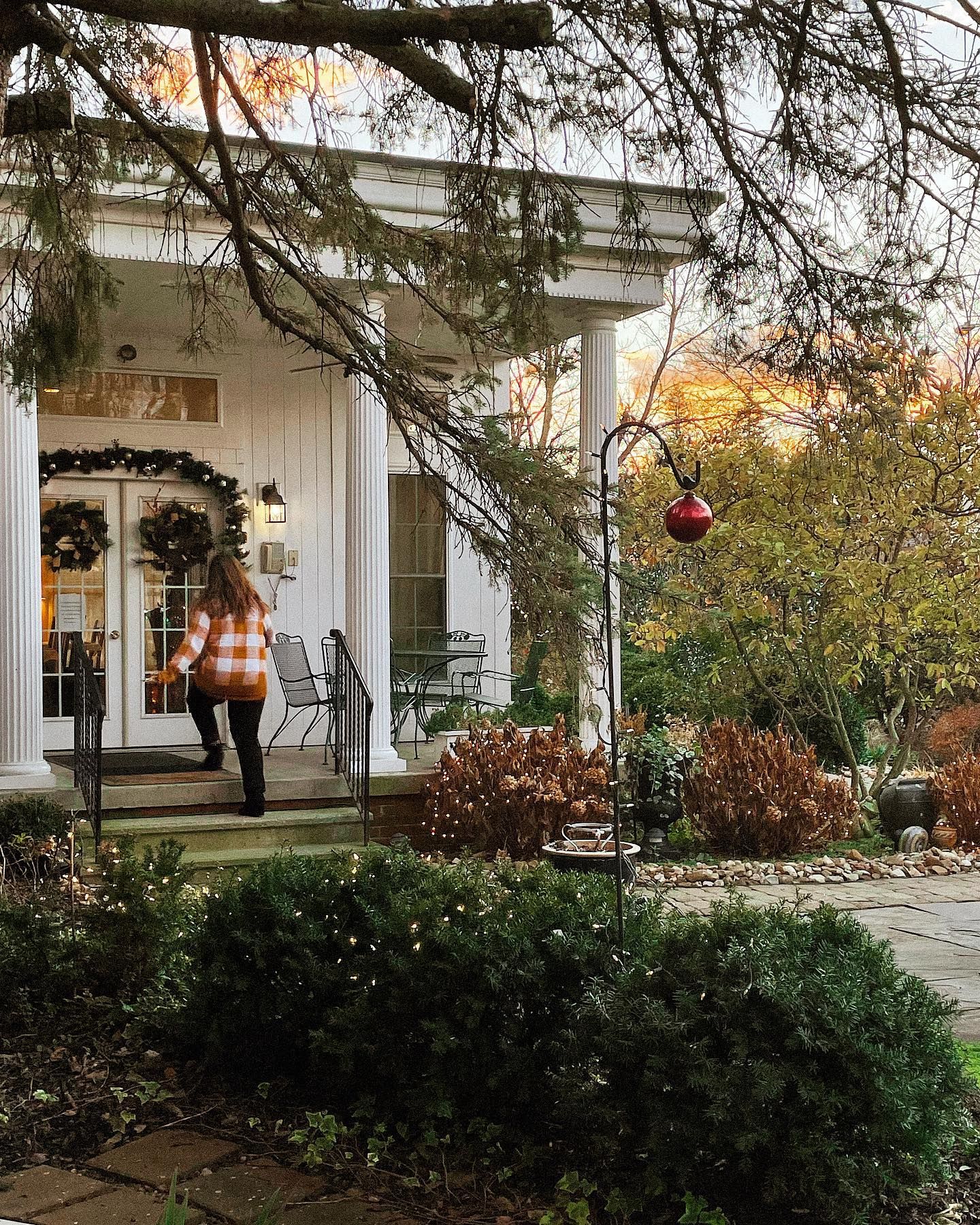 Woman enters a white porch with wreaths on the door, surrounded by greenery.