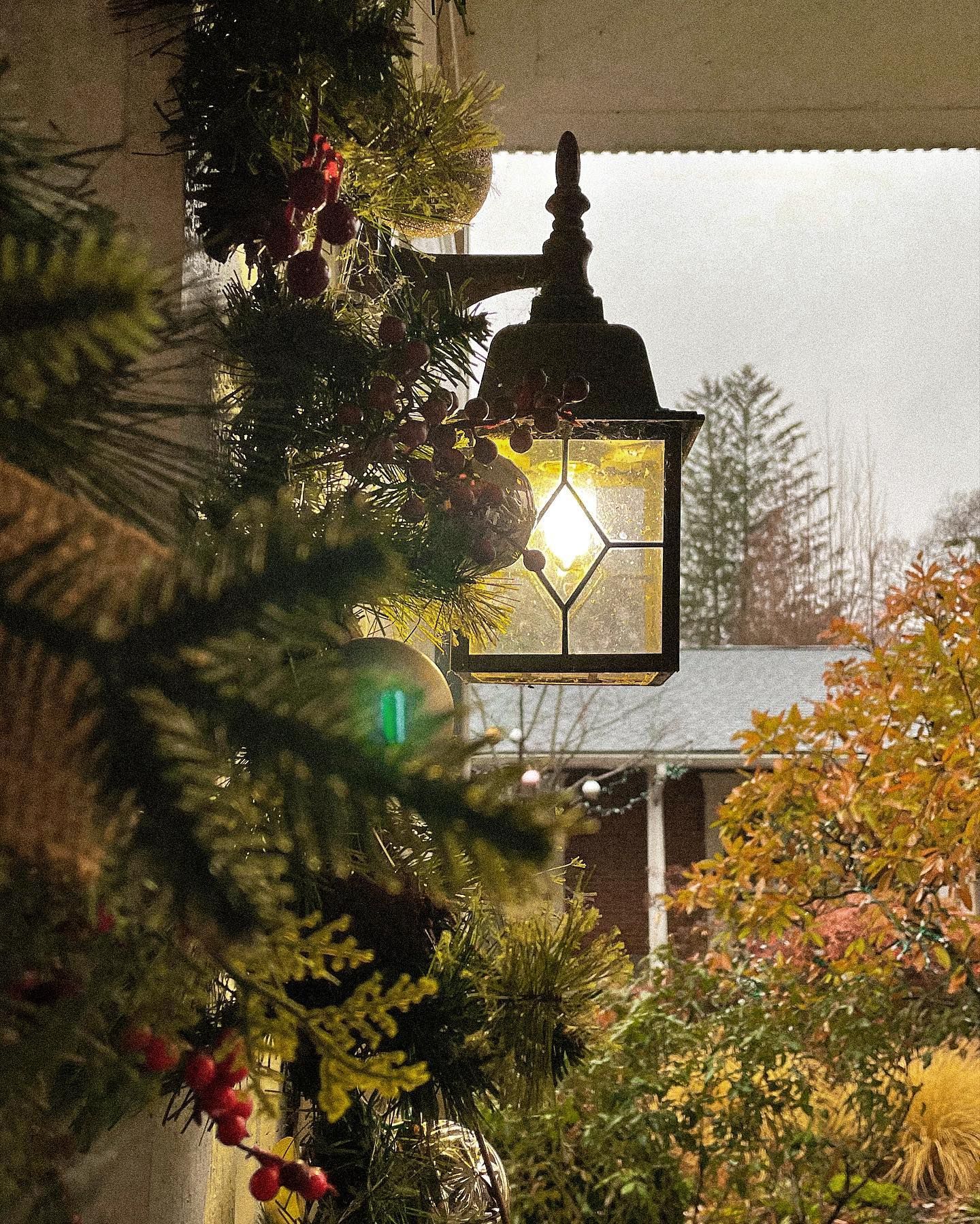 Outdoor lantern with lit bulb, framed by Christmas greenery and autumn foliage.