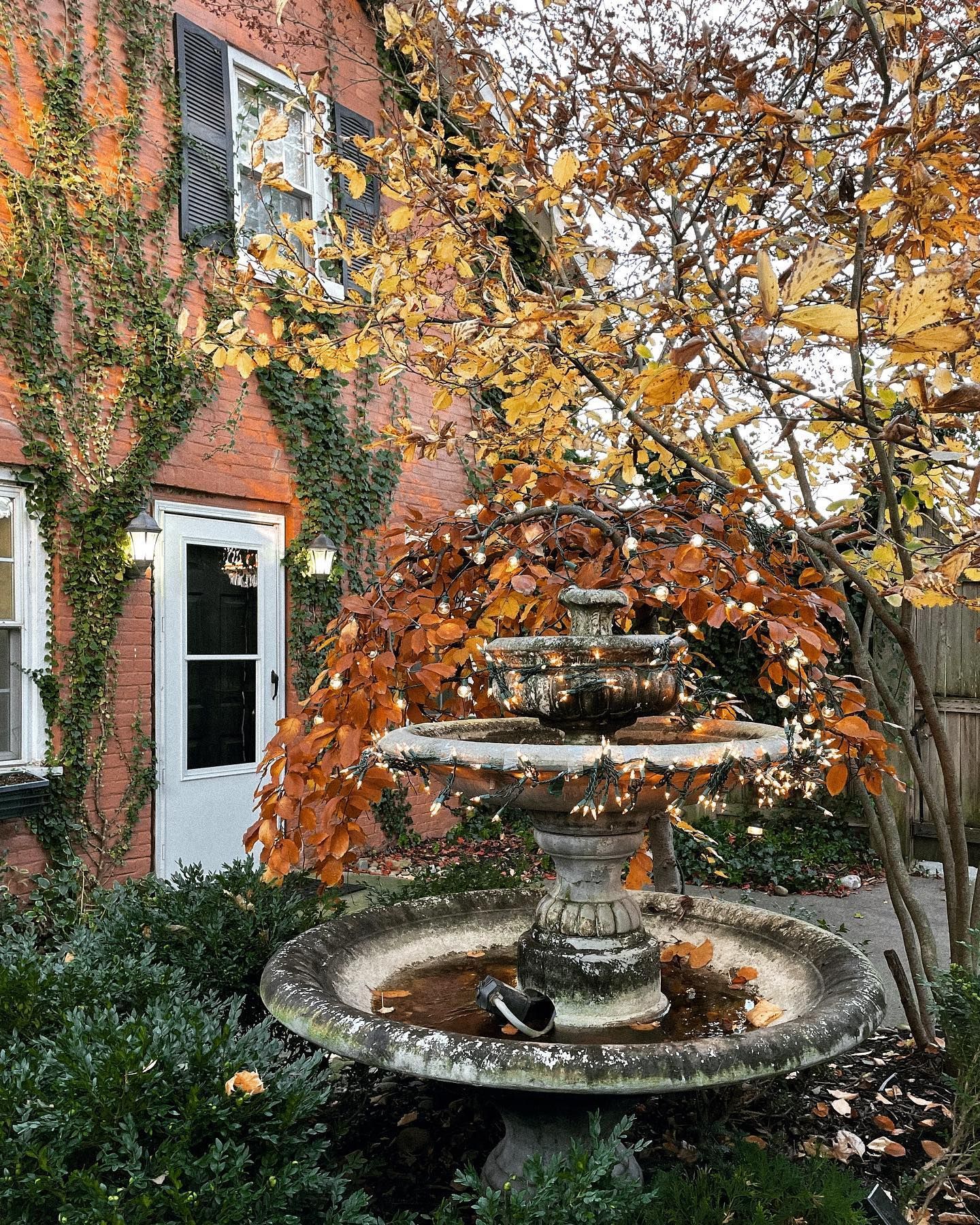 Stone fountain in a garden with autumn leaves and red brick building.